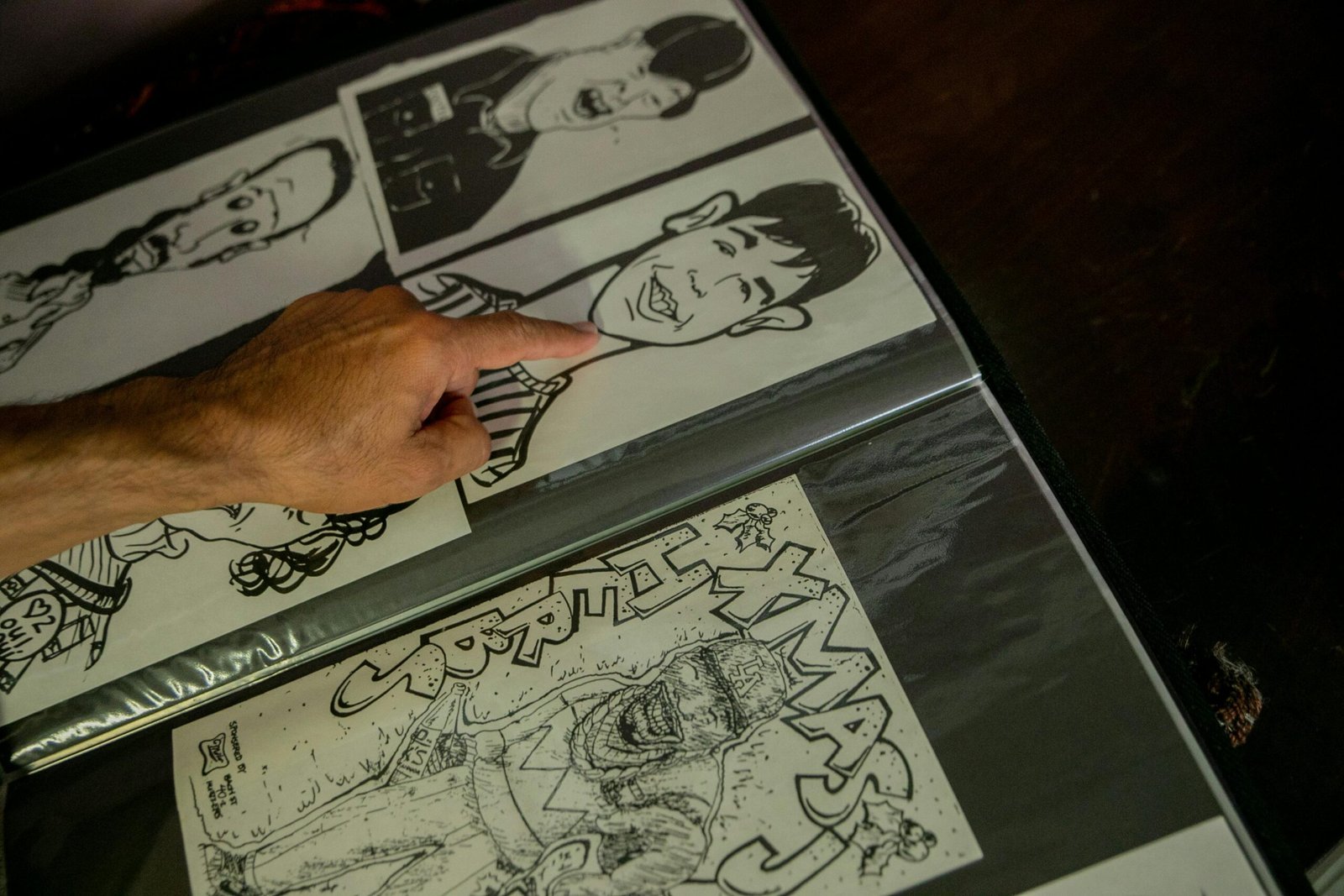 Close-up of a hand pointing at caricature sketches in an art portfolio on a dark surface.