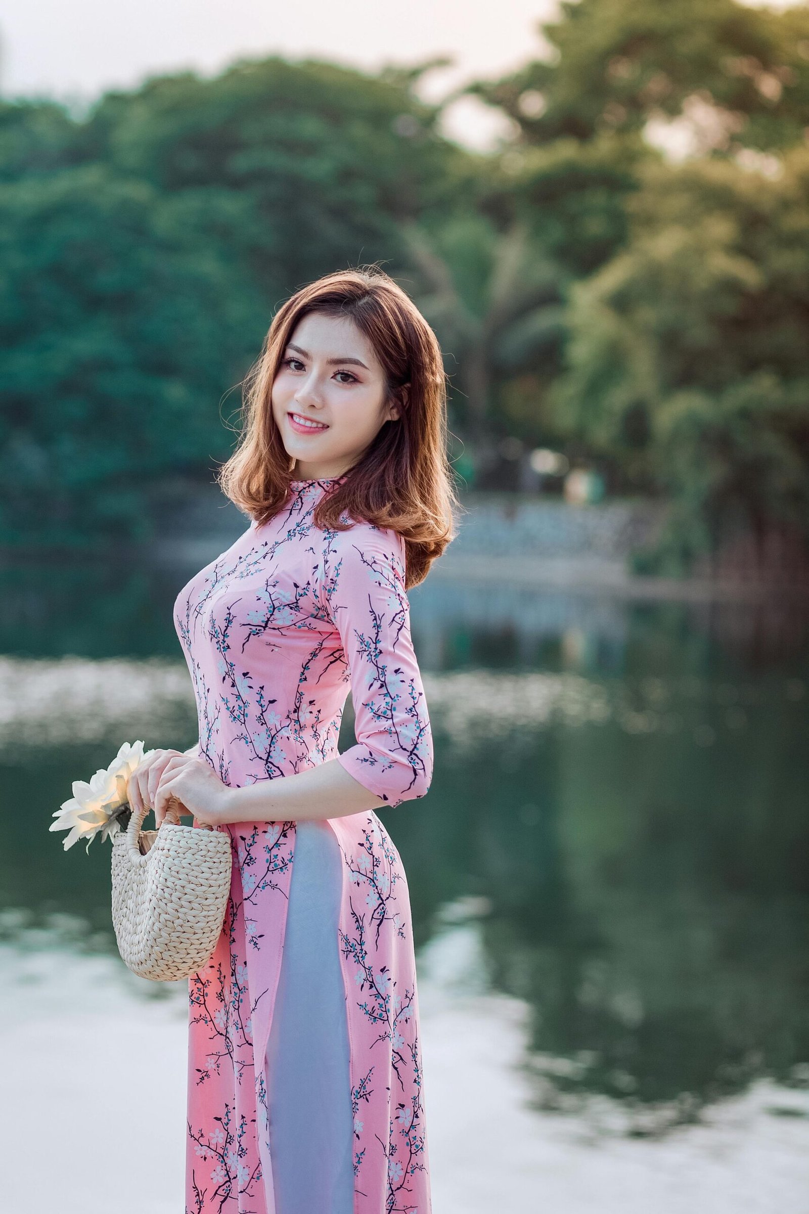 A smiling woman poses gracefully in a pink dress by a serene lakeside, holding a woven basket.