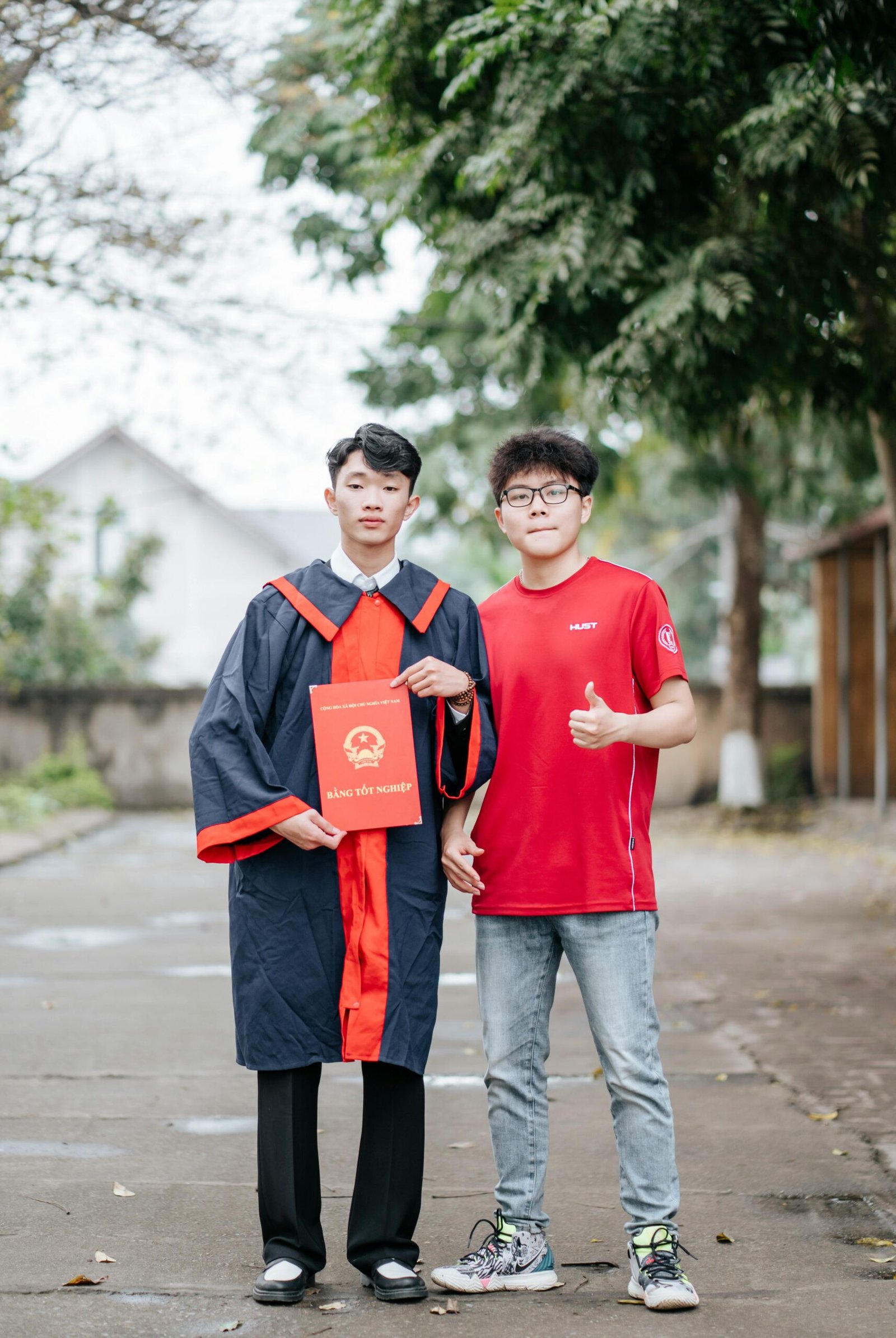 Two friends celebrate graduation outdoors, one in cap and gown holding a diploma.