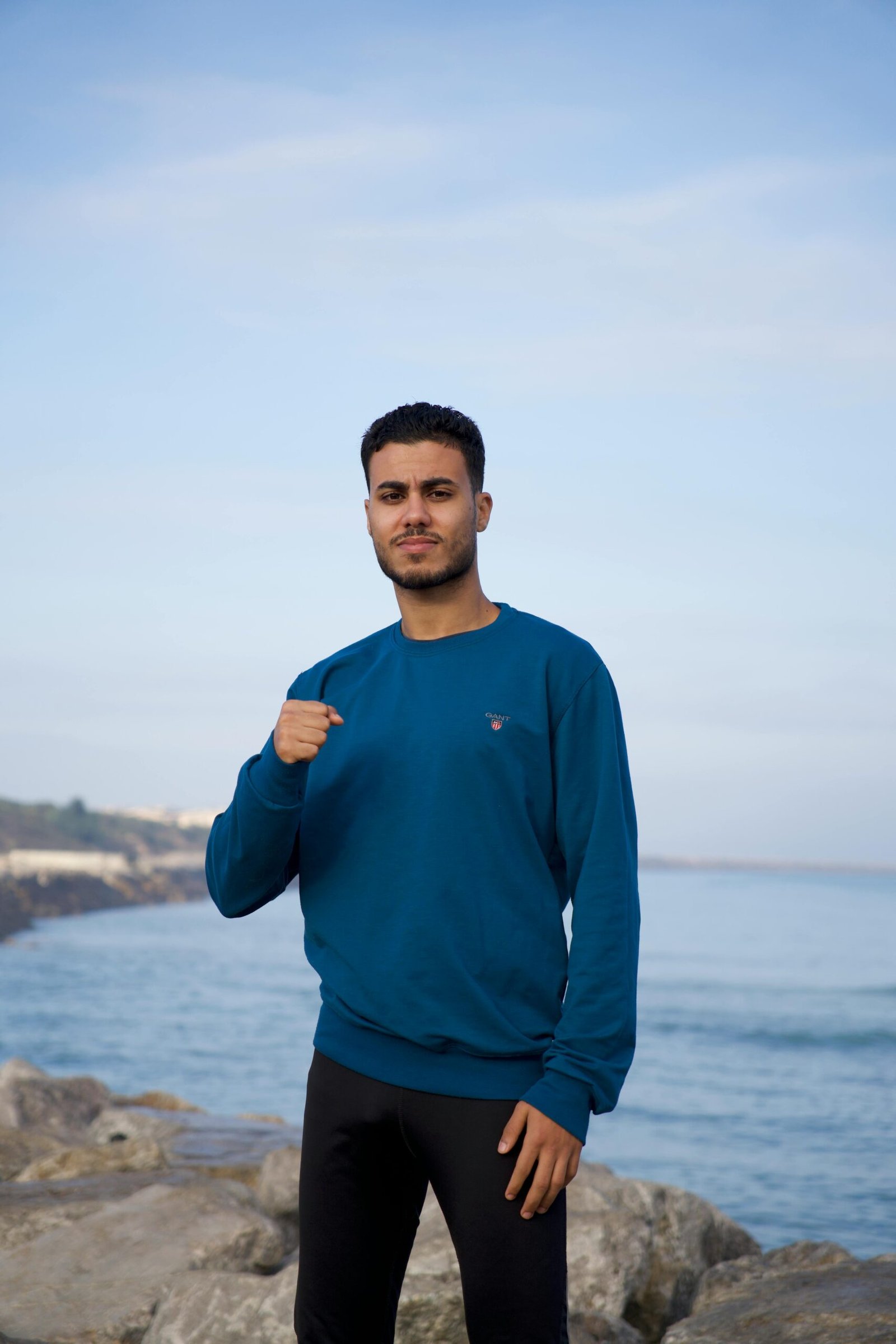 Young man in blue sweatshirt posing on a rocky beach with ocean view.