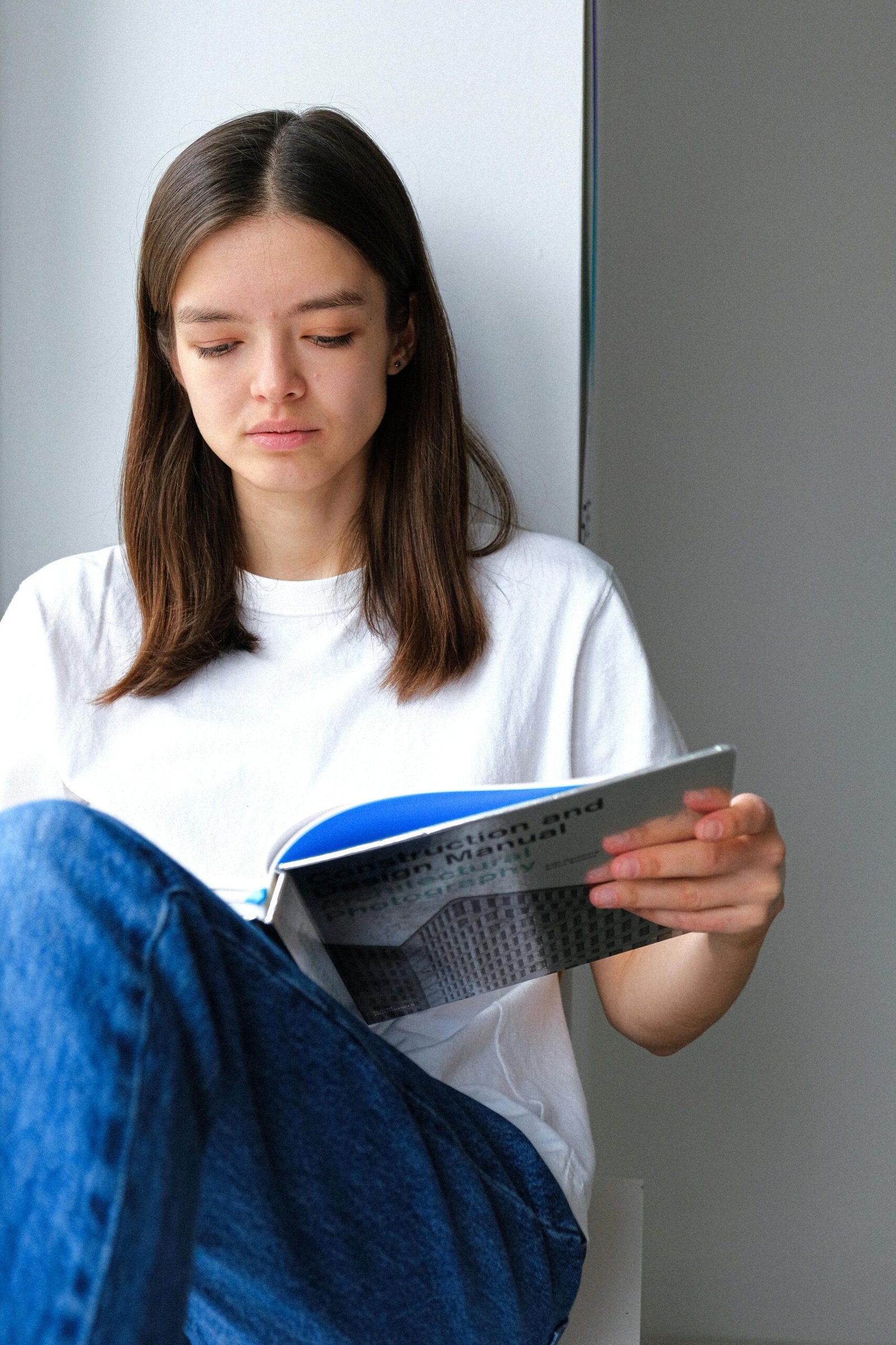Young woman in a casual setting, deeply engrossed in reading a book by a window at home.