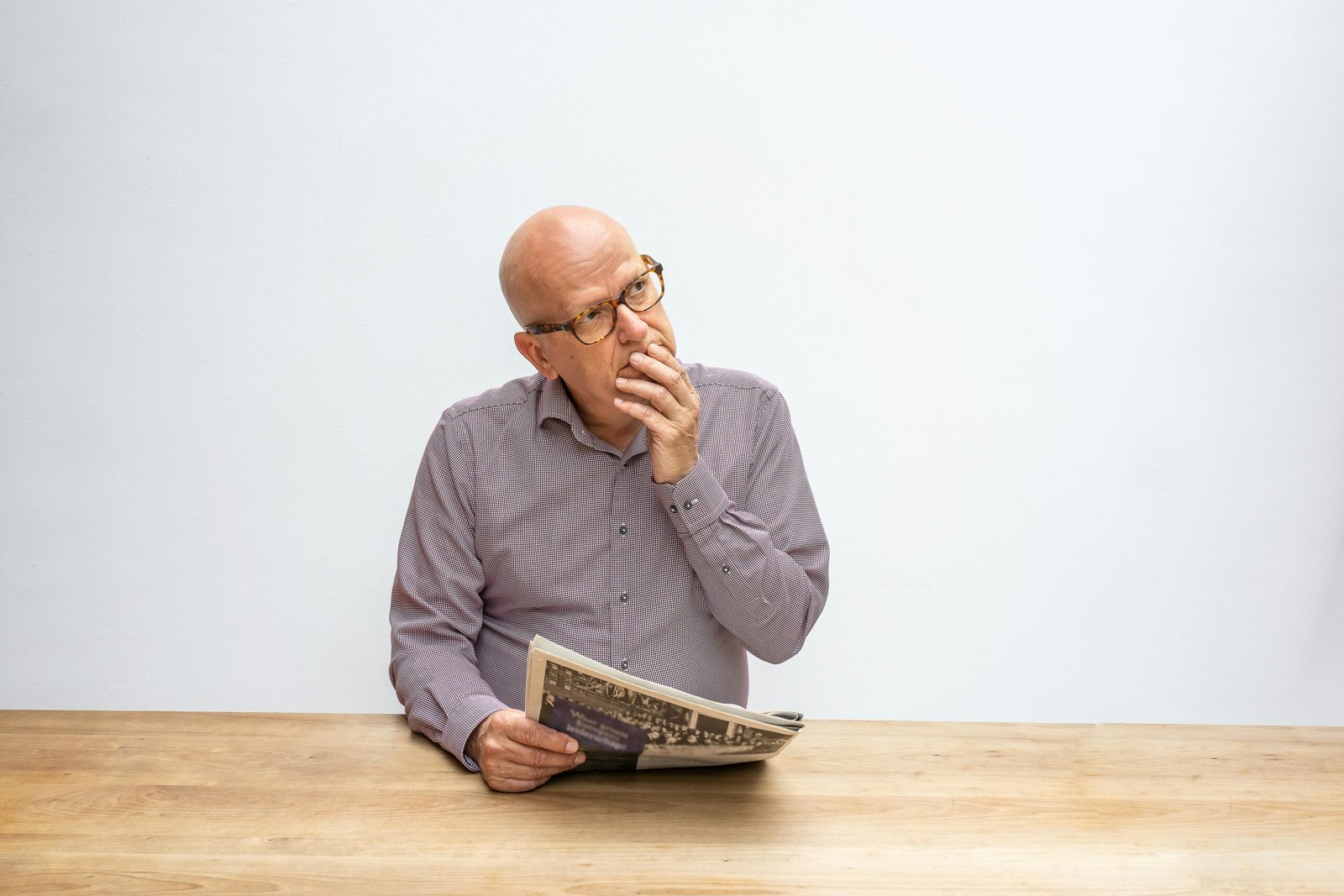 Bald senior man with eyeglasses thinking while holding a newspaper at a wooden table.
