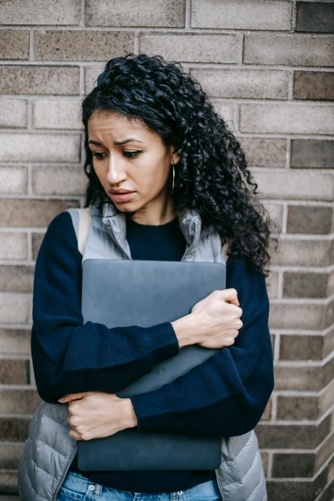 Tired Hispanic female with curly hair leaning on brick wall while carrying netbook and looking down